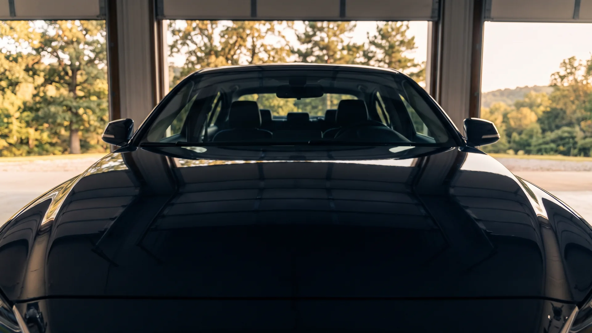 The clean, freshly coated hood of a dark car standing in an open workshop doorway, with an Arkansas treeline in the daylight beyond it.