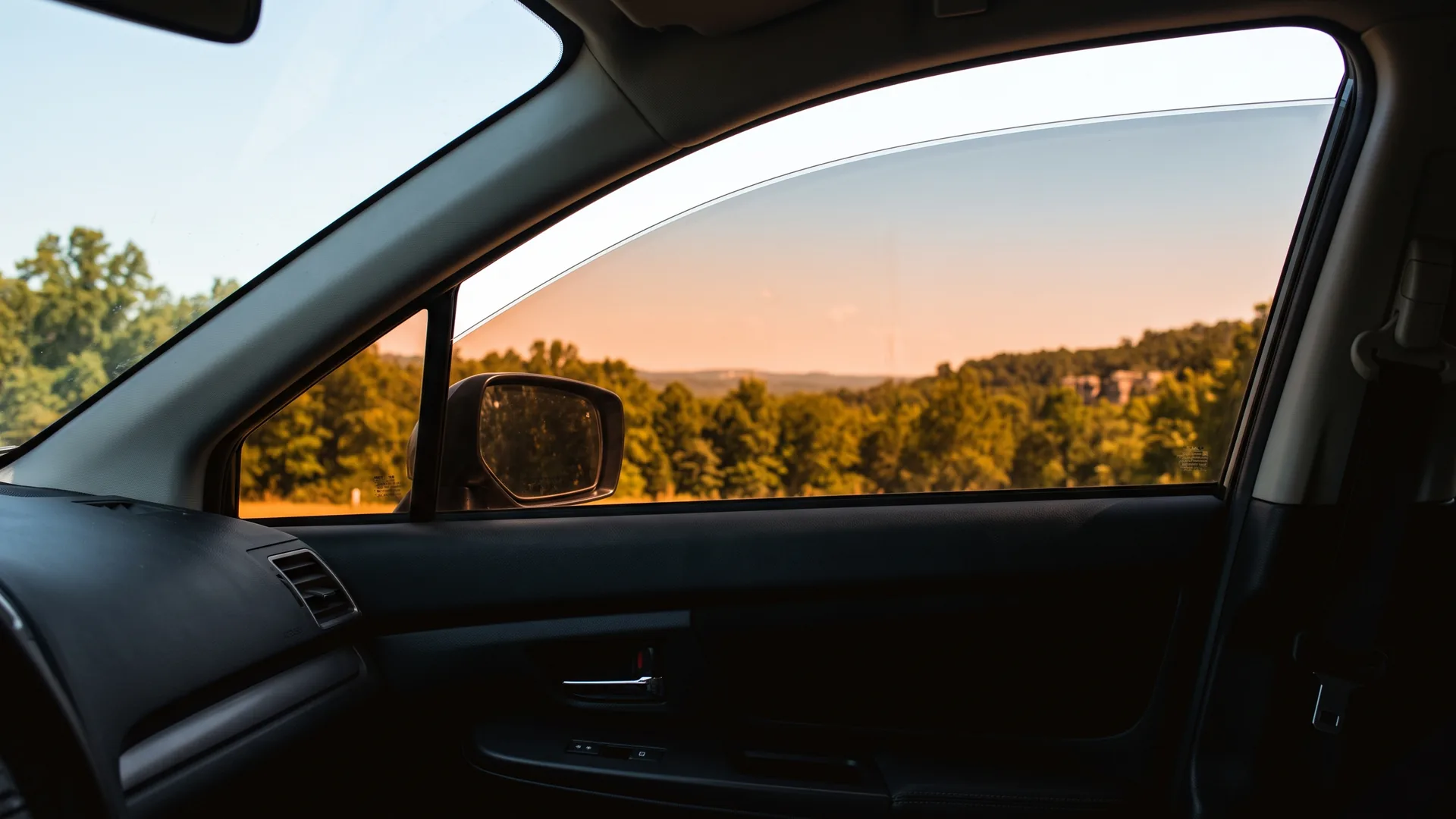 The view out through a tinted front side window on a hot afternoon. An Arkansas hardwood treeline and low hills are clearly visible through the glass, while the cabin side of it sits in shade.