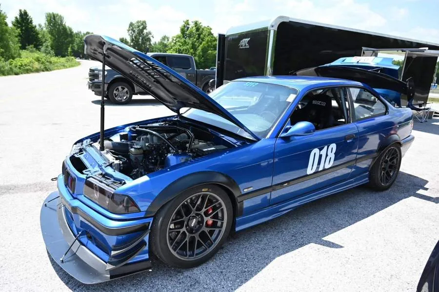Blue BMW E36 track car numbered 018 with the bonnet propped open, beside a race trailer at a circuit.