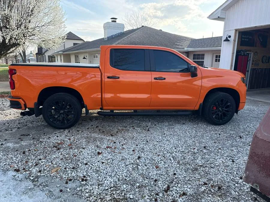 Orange Chevrolet Silverado crew cab with tinted windows and black wheels, parked on a gravel drive.